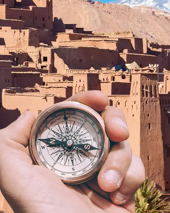 A hand holding a compass in front of the earthen kasbah of Aït Benhaddou