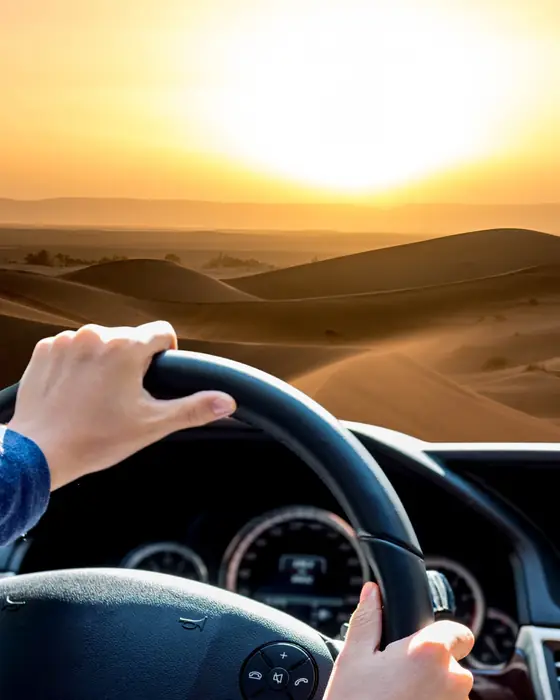Hands on a steering wheel, driving towards sand dunes and a low sun