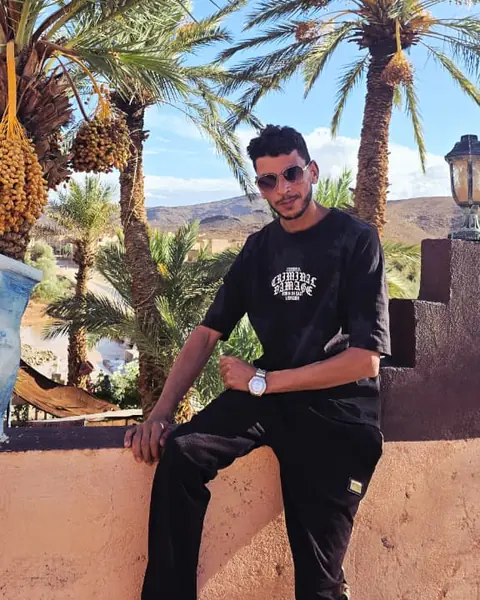 Mohammed sitting on a sunlit wall under date palms heavy with fruit, with desert hills behind him
