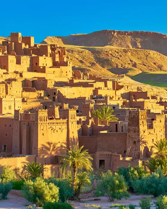 The earth-built kasbah of Aït Benhaddou near Ouarzazate in warm light, with palms below and hills behind