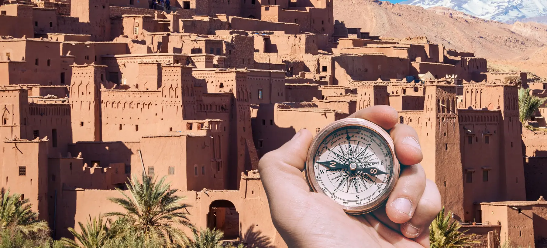 A hand holding a compass in front of the kasbah of Aït Benhaddou, with snow on the High Atlas behind