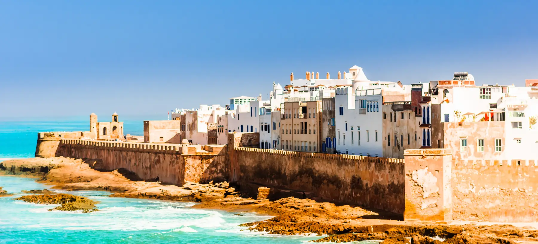 The ramparts and white houses of the old city of Essaouira above turquoise water and rocks